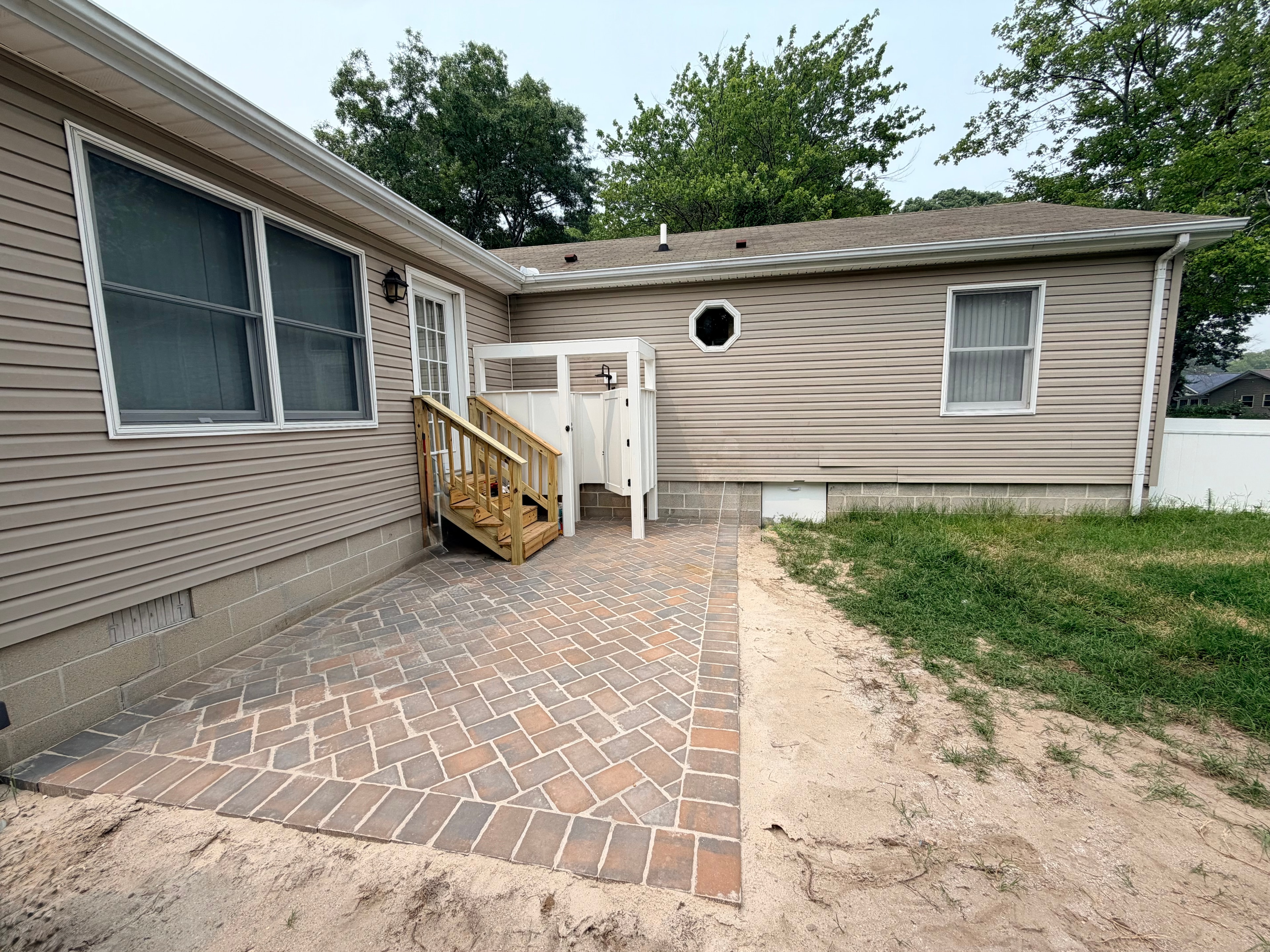 Completed brick paver patio with wood deck steps and outdoor shower enclosure on residential home